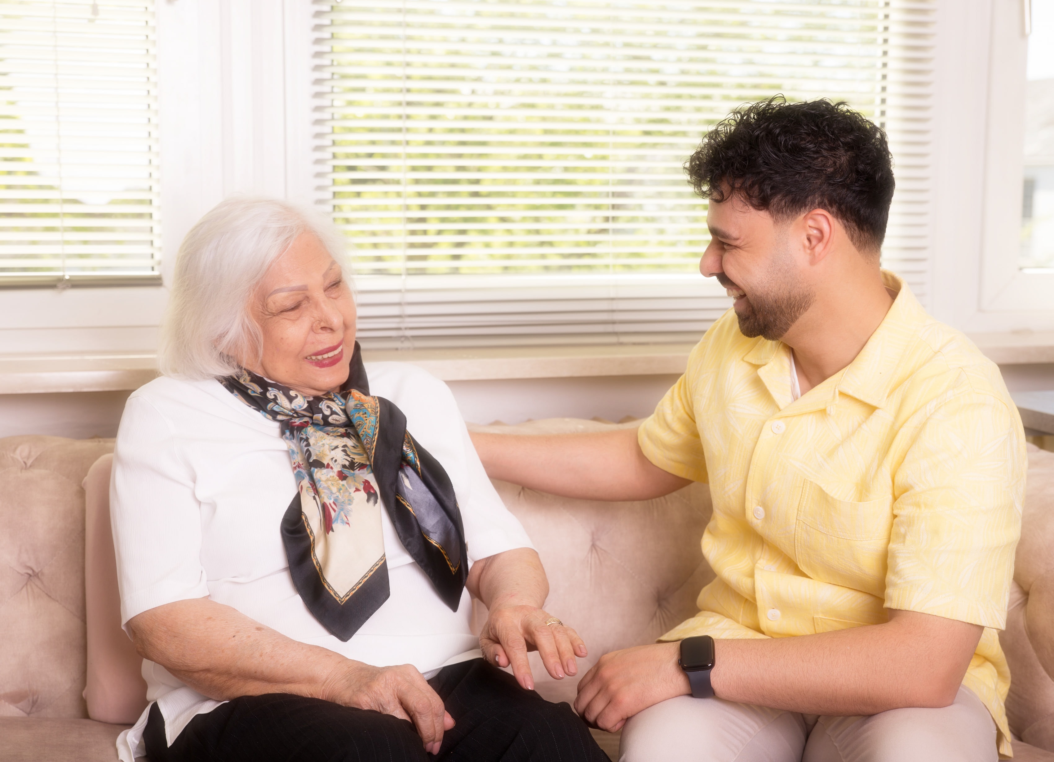 Ein Pflegefachmann und eine Seniorin lachen gemeinsam auf dem Sofa in ihrem Zuhause